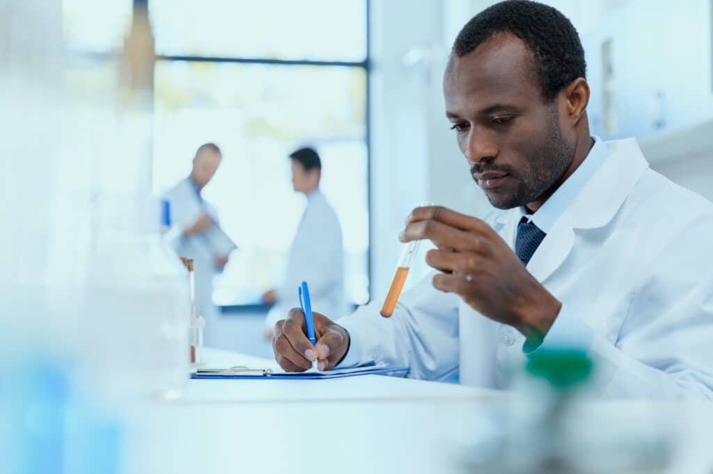 African american scientist in white coat holding and examining test tube with reagent, laboratory